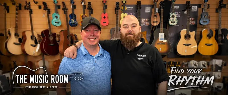 Two men smiling in front of The Music Room YMM's wall of acoustic guitars and ukuleles